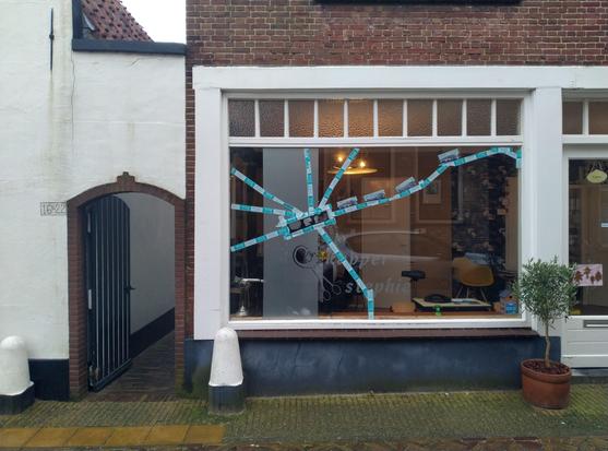 Photo of a broken window of the shop of a hairdresser, fixed with blue tape in a starlike pattern. Next to the window the access to an alleyway through a white gate with an open door.