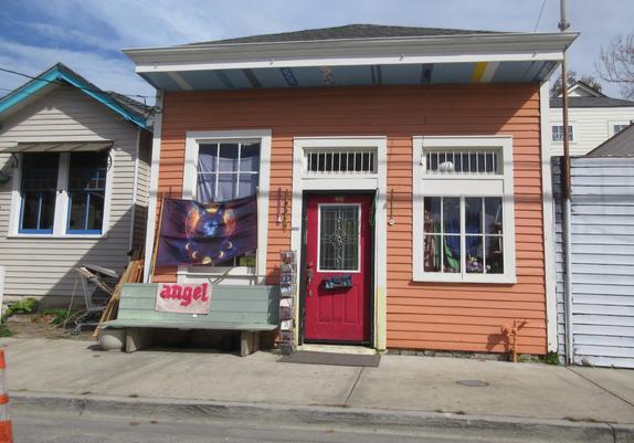 Urban street, concrete sidewalk, with close-set single story buildings.  Central building is a "shotgun" style wooden late 19th century vernacular building used as a shop. Windows are on either side of the central doorway. The front is painted orange with white trim on the window and door frames, the door is painted red.  The door has a window with geometric patterns in the glass, below which a metal mail box hangs crookedly. 

To left of the door, the window is partially covered with a banner depicting a wolf and the phases of the moon.  Below it is a wooden bench on the sidewalk, with a towel or mat with the text "angel".  Between the bench and the door is a small metal wire rack holding compact discs for sale.