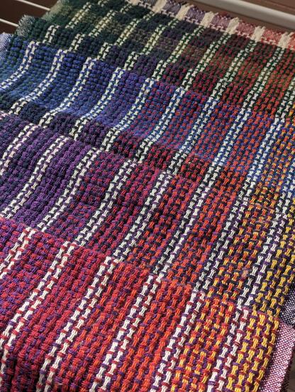 handwoven huck fabric in a rainbow gradient laid on a drying rack. the fabric transitions from reddish purple mixed with yellow in the bottom right corner to dark green in the top left