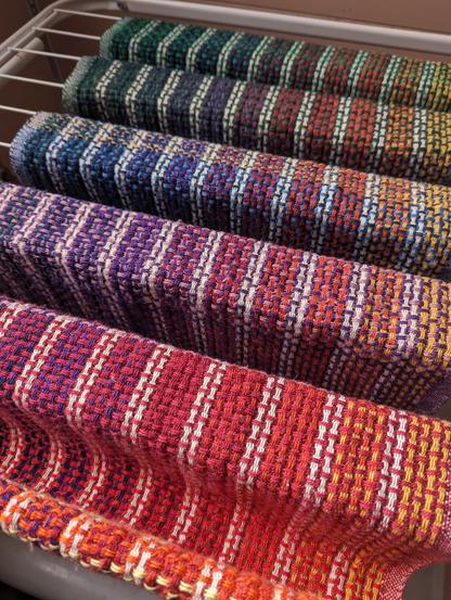 handwoven huck fabric in a rainbow gradient laid on a drying rack. Rainbow and white stripes travel vertically throughout the cloth, intersected by stripes of brighter and contrasting colors
