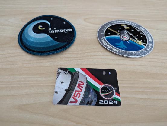 Due toppe di stoffa circolari e una tessera di plastica con la forma e le dimensioni di una carta di credito sono poggiate su una superficie di legno chiaro. Le toppe sono gli emblemi di missioni spaziali, l'immagine sulla tessera mostra la Terra e la Lune riprese dallo spazio con una capsula Orion in primo piano.