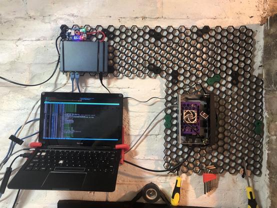 Image of a white wall with electronics mounted onto a black plastic frame made of hexagons.
Bottom left is a laptop displaying  htop, above the laptop is a 8 port network switch and it’s new power supply and then on the right is an orangepi sbc. There are various cables around