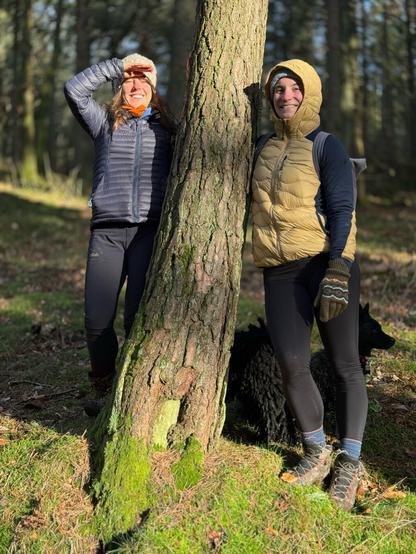 Two ladies standing beside a tree on a sunny Winter’s day. The weathered bark is covered in moss. The forest behind them has the late afternoon sun filtering through the trees.