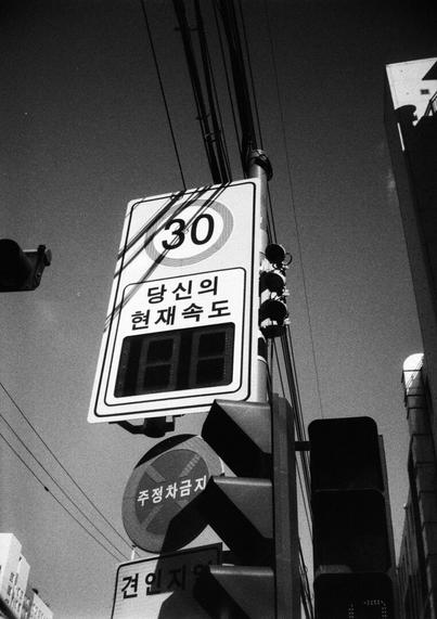 Vertical and black and white photo. There is a vertical rectangular sign in the middle of the photo. There is an LED window that says 30 in the circle at the top and "Your Current Speed" at the bottom and up to 100 but it is now off. You can see the side of the traffic light below and behind it are "No parking or stopping" and "Tugging" signs. Wires are running densely in the vertical direction over the signpost.