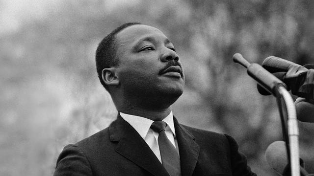 An upper-body photo of Martin Luther King Jr. He is standing before a bank of microphones, and looking out upon an audience.