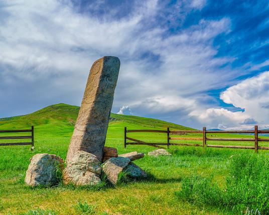 A thin leaning stone about two meters tall, carved with stylized figures. The scene is set on green rolling hills with dramatic clouds in a deep blue sky.