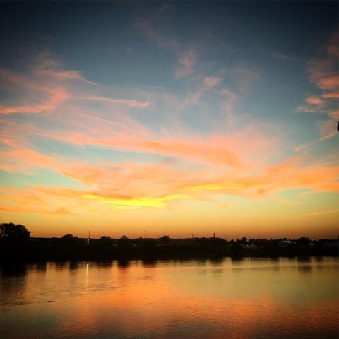Sunset sky with orange and blue clouds reflected in a body of water with a silhouette of a distant shoreline across the Arkansas River, Tulsa, Oaklahoma.