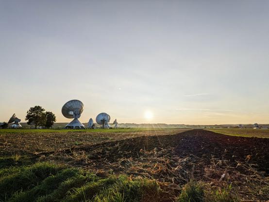 Blick über ein Feld Richtung untergehende Sonne. Im Bild sind diverse große Satellitenantennen der Erdfunkstelle Raisting.