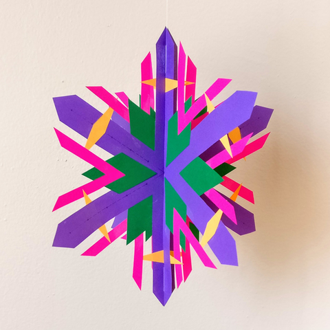 A purple, pink, green, and orange three-dimensional paper snowflake hangs from a thread in front of a beige wall.