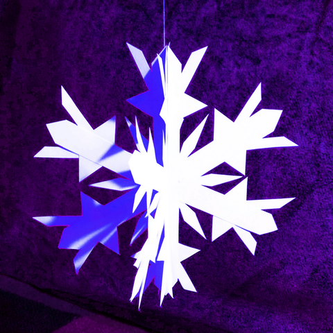 A white three-dimensional paper snowflake hangs from a thread in front of a purple velvety background.