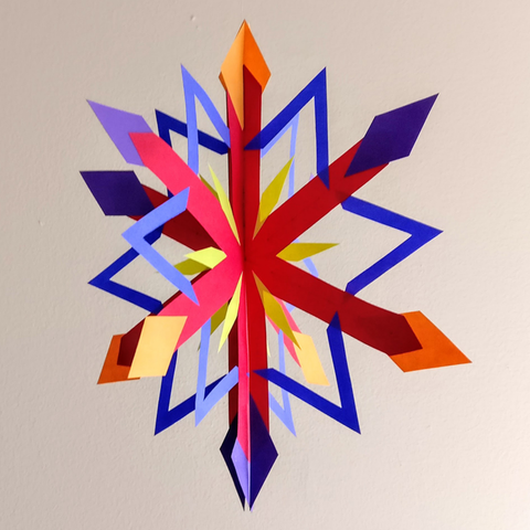 A red, blue, orange, and yellow three-dimensional paper snowflake hangs from a thread in front of a beige wall.