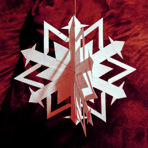 A white three-dimensional paper snowflake hangs from a thread in front of a red velvety background.