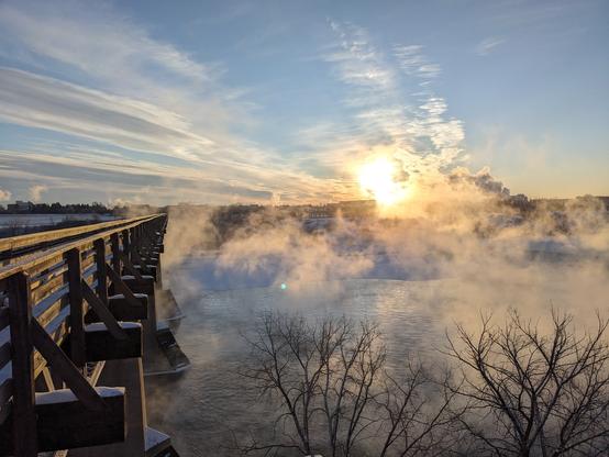A bridge over an icy river with water vapor rising. Blue sky with a low sun.