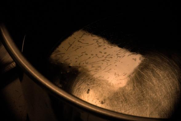 Rectangle of reflected light (kitchen window) on the surface of a calm pot of water, filligreed with swirls of oil.  A rough circle of abraded steel on the base of the pot is below the rectangle.The curved edge of the pot frames the scene.