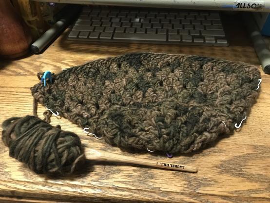 An half-crocheted  beanie/toque hat, in variegated shades of brown & dark brown, lies on an oak desk, next to a wooden crochet hook stuck into a rather small ovoid ball of the yarn in use. A few white & blue stitch markers indicate the center line of the hat; and a keyboard & monitor stand lurk in deep shadow at top of frame.