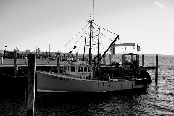 A black and white photograph of a small commercial fishing boat at dock. The name “Janet Maria” is painted on the bow.