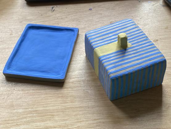 a photo of a leather hard butter dish with the tray to the left and the lid part to the right. I painted both with blue underglaze, the upper side of the tray and the surface of the lid. The lid is done in blue stripes with the handle in yellow (IKEA vibes, totally). Now the handle not only looks like a stick of butter, it is a melting stick of butter and the yellow is flowing down one side of the lid. It looks pretty terrible, but this is what I have to work with from know because this is on me, trying to be funny.