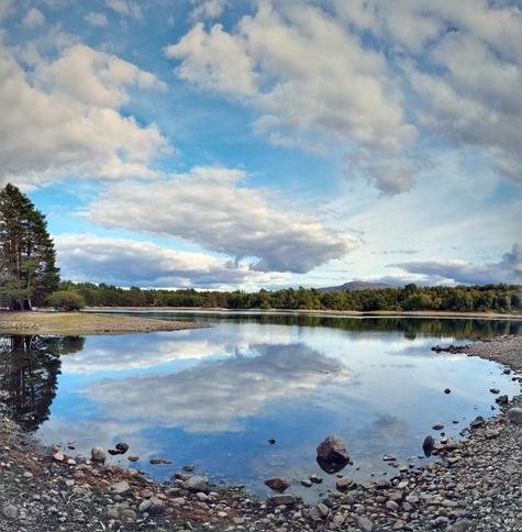 A view over Loch Vaa. The water is still and like a mirror, reflecting the blue sky and clouds floating gently past. On the far shore fairly dense trees can be seen, The water level is low, showing the rocky shore that would normally be a bit more hidden.