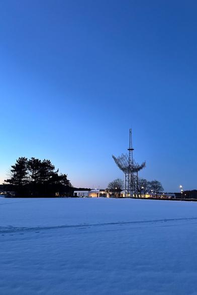 Snow-covered field with trees on the left, a distinctive tower structure in the center, and buildings with lit windows in the background, under a twilight sky.