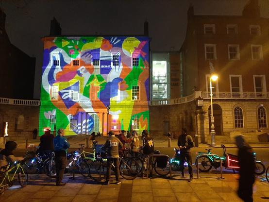 Image shows people with bicycles gathered opposite the Hugh Lane Gallery. Some bikes are light up with fancy lights. The gallery in the background is illuminated with a projection of a Claire Prouvost Illustration which looks great in the darkness.