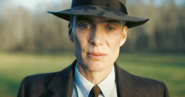 A still from the film Oppenheimer. A blue-eyed white man with a sad expression, wearing a grey jacket and hat, looks towards the camera, with a grassy field behind him.
