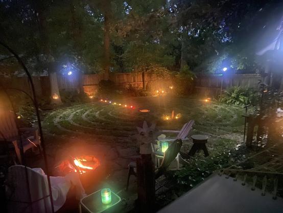 A night-time back yard scene with a 7-circuit labyrinth lit with candles, including seven chakra candles along the western axis. In the foreground is a fire pit with a fire going in it.