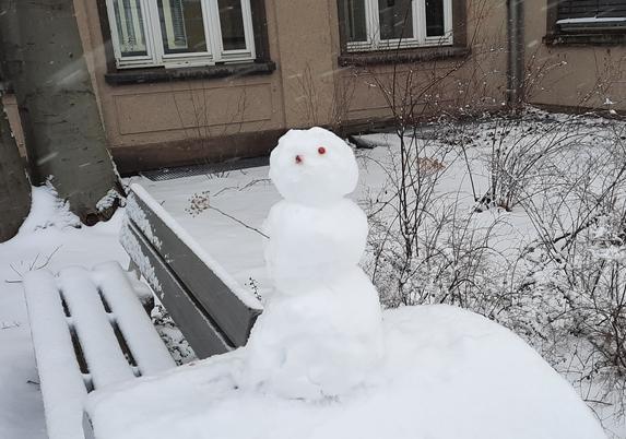 Kleiner Schneemann vor einer Bank und einem Gebäude, mit 2 Augen und 2 kleinen Zweige als Arme, die sich vorne feingliedrig wie Hände und Finger verzweigen. Die Haltung vermittelt die Botschaft des freudigen Willkommens.