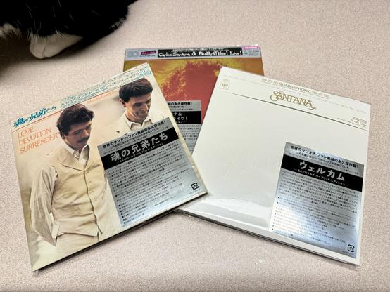 Three Hybrid Super Audio CDs by Santana are lying on a countertop.  A tuxedo cat’s left paw can be seen in the background
