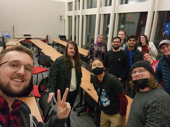 Several people are standing clustered together in a university classroom, posing for a selfie together.