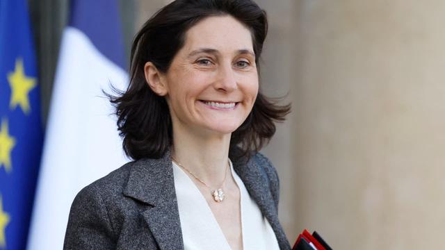 Une femme maigre sort d'un bâtiment de la République : drapeaux européen et français sont à gauche.

Elle sourit à un·e des journalistes à droite de la caméra.
Veste en tweed gris, collier discret, cheveux bruns coupés au ras des épaules), pas de boucles d'oreilles apparents. Les lèvres horizontales et les plis du sourire barrent le visage en macron. [Le macron ‹ ◌̄ › est un diacritique de plusieurs alphabets : latin, grec et cyrillique. Il prend la forme d'une barre horizontale. Son principal rôle est d'indiquer que le signe qui le porte reçoit une quantité vocalique longue.]

La ministre française des Sports, Amélie Oudea-Castera, quitte le Palais présidentiel de l'Élysée après la réunion hebdomadaire du cabinet, le 20 décembre 2023. - LUDOVIC MARIN, AFP