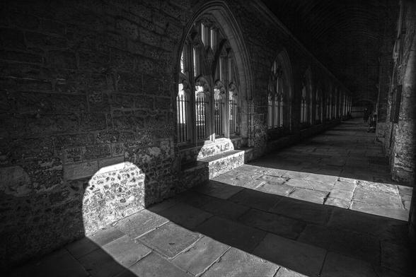 A black and white photo with fairly high contrast lending a lot of depth to the shadows. We're standing in medieval cloisters, against the right hand wall and looking down the length of the walled-in area of this religious building as it stretches diagonally from bottom left up to the right. The long wall we're looking at is punctuated by large gothic windows, each at least two metres across at the base, and over that in height, and each broken up into smaller, tall gothic-arched window sections framed with stone columns: four along the bottom, two sets of two smaller above, then a single set of two at the high point of the main frame. The walls comprise smaller stones set in concrete at the bottom, topped with larger blocks of stone above. Through the nearest arch we can see a tiny fraction of the grounds beyond. Light comes from two open doorways in the wall to our right. We can't see the doorways but can see the shafts of sunlight that stretch across the flagstones on the floor and hit the far wall at an angle, almost appearing as bright gravestone markers against the dark surroundings. Deep shadows hide the ceiling from view and the far end of the cloisters is lost in blackness. It feels cool, shaded, and quiet here.