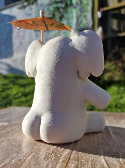 Clay model of the mastodon elephant mascot sitting down and holding a cocktail umbrella. The photo is taken from behind and he had rotund, pert buttocks