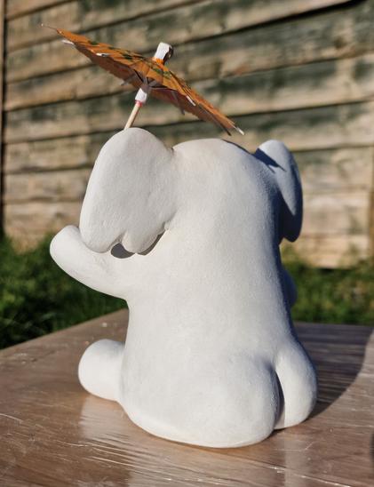 Clay model of the mastodon elephant mascot sitting down and holding a cocktail umbrella. The photo is taken from behind and he had rotund, pert buttocks