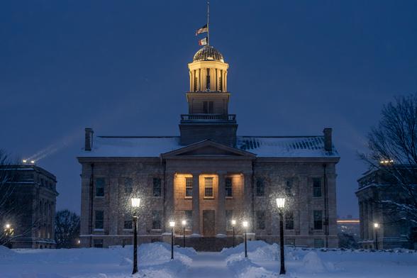 Image of the old Iowa State Capitol Building on the campus of the University of Iowa in Iowa City, Iowa. The image was taken at night during a heavy snow shower. The capitol building is centered in the frame with portions of similar style buildings visible on either side. The University of Iowa refers to the architectural style as Greek revivalist. The architect was John Francis Rague. A sidewalk in the lower frame leads directly to the building's stairs and entry way and is lighted with streetlights that have black poles and lantern-like lamps. The streetlights highlight portions of the sidewalk leaving the area between lights in shadow. The lights are somewhat blurred due to the heavy snow falling past the. The capitol building is in shadow except for its entrance which is highlighted by lights that are directed downward from behind a neoclassical facade and the capitol dome which is highlighted by large lights mounted on the roofs of the nearby buildings. The predominant colors are a dark blue-grey that extends from the bottom of the frame to the overcast sky above, white light coming from the streetlights and building roofs, and a warm golden light at the building's entrance and dome.