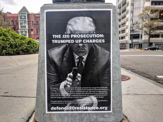 A poster wheatpasted to the base of a light pole in downtown DC reading "The J20 Prosecution: Trumped up charges" with a picture of Donald Trump brandishing a gun and the website defendJ20resistance.org.