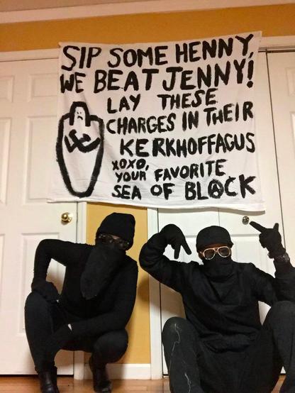 Two people in full black bloc pose with a banner reading "Sip some henny, we beat Jenny! Lay these charges in their Kerkhoffagus! xoxo, your favorite sea of black" after the defeat of despicable prosecutor Jennifer Kerkhoff in court.