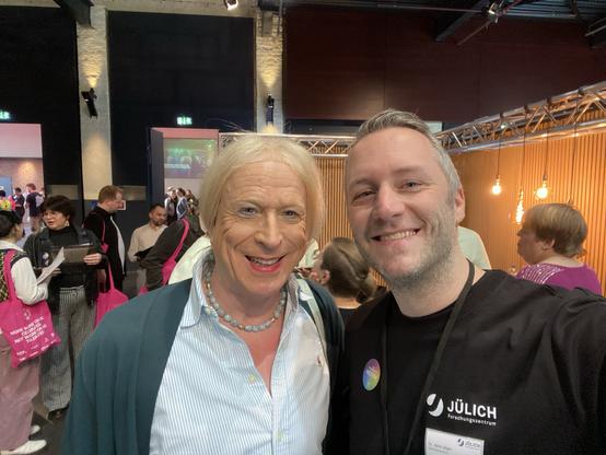 Selfie mit Georgine Kellermann auf der Messe, sie in Bluse und Jacke, ich im FZJ-T-Shirt; im Hintergrund Messestände und Menschen mit rosa Taschen.