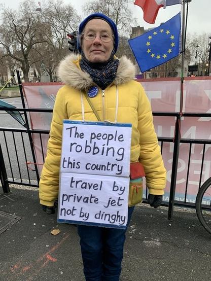 Woman wearing a yellow jacket standing in front of a railing with an EU flag wearing a poster which says "That people robbing this country travel by private jet not by dinghy."