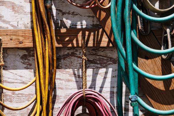 A color photograph showing detail of an open workshed door. The door itself, made from plywood coated in fading white paint, is the background. On it, the unpainted wooden cross and diagonal braces are visible.

Hanging from nails and supported by ropes, on the braces, portions of other items are visible. To the left, a heavy duty yellow electrical extension cord is visible, looped multiple times, from top to bottom of the frame. The top of a coiled red extension cord is partially visible at the bottom center, supported by a rope attached to a nail, mid-frame. A second similar cord is visible at the top edge above it. To the right, a portion of a coiled green water hose is visible.