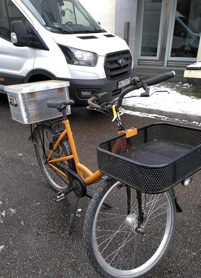 Ein Lastenrad steht auf der Straße. Dahinter ein Sprinter. Das Fahrrad hat vorn einen Aufbau zum Transport von Gegenständen und hinten eine Box.