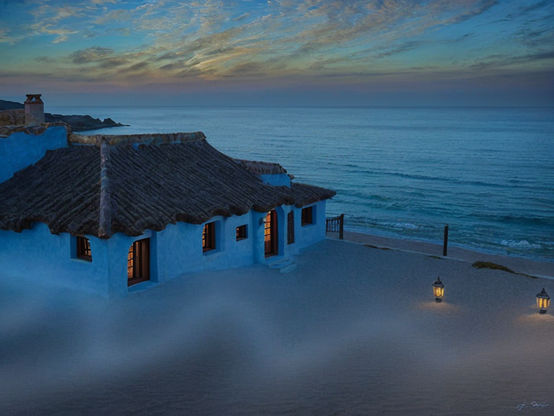 A beach house at sunset, shrouded in a soft mist, with warmly lit windows. Two lanterns light the beach.