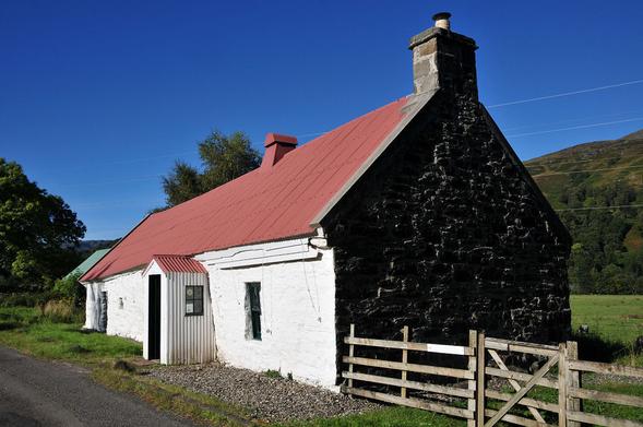 The Moirlanich Longhouse. The image shows a high contrast oblique view of a cottage with a stone gable on the right and a white-painted front on the left and a dull red corrugated iron roof. There's part of a road in the left foreground. The sky is a bright dark blue.