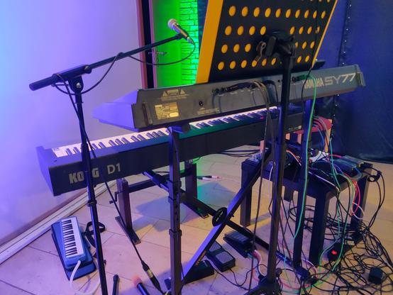 Keyboard-Setup auf der Bühne, von hinten: auf dem Ständer ein Yamaha SY77 Synthesizer, darunter ein Korg D1 Digitalpiano, auf dem Fußboden eine blaue Melodika. Notenpult und Mikrofonständer stehen hinter bzw. neben den Keyboards.