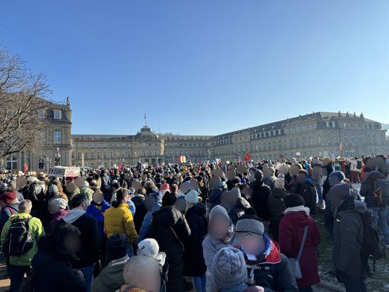 Eine Masse von Menschen, teils mit Demoschildern, Transparenten oder Fahnen vor dem neuen Schloss in Stuttgart. Darüber blauer Himmel.