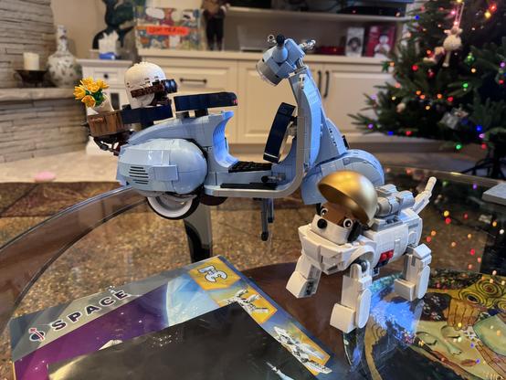 Lego Vespa in background with basket of flowers on back and helment perched in front of that. Right, Lego dog astronaut.