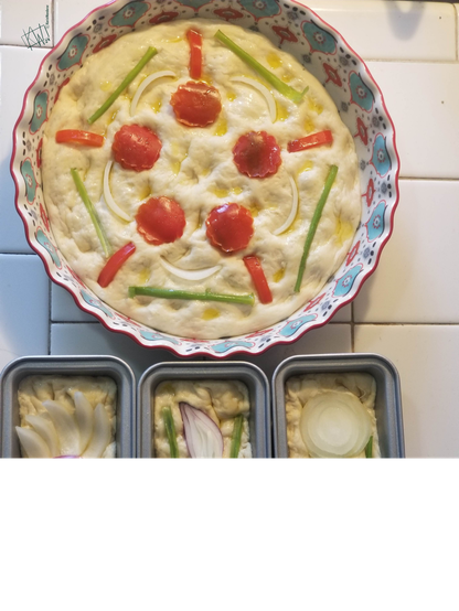 1 round & 4 small loaves of focaccia, decorated with produce & ready to go in the oven. The round one has a mandala made with red peppers & red & green onions. The rectangular ones have a sliced onions arranged to look like flowers.