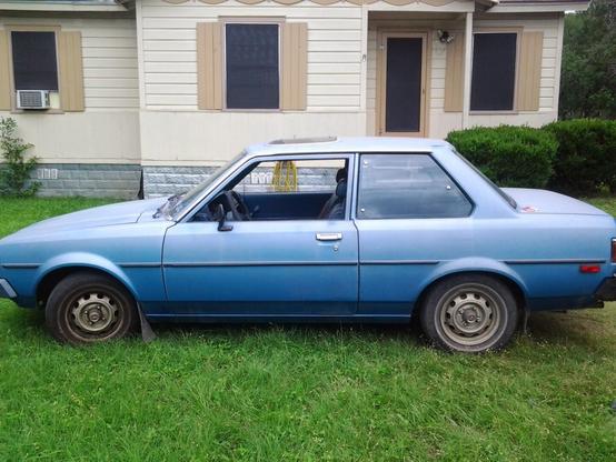A classic blue Toyota Corolla parked in front of a small cream colored home. The car is sitting underneath a tree.
