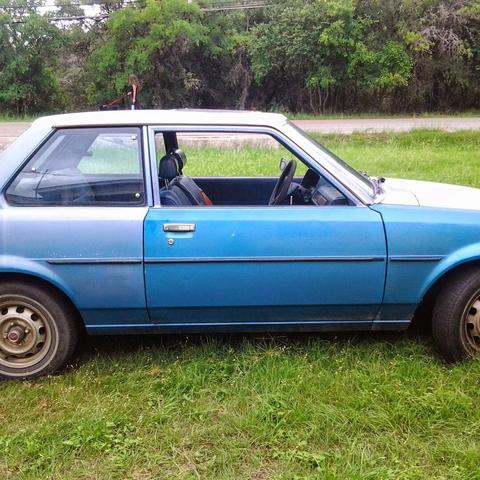 A classic blue Toyota Corolla with semi-faded paint. It is parked in a vibrant patch of green grass. The driver's seat has a tear in it and has blue interior. The car is gleaming in the sunlight as a person with their dog is walking in the background.