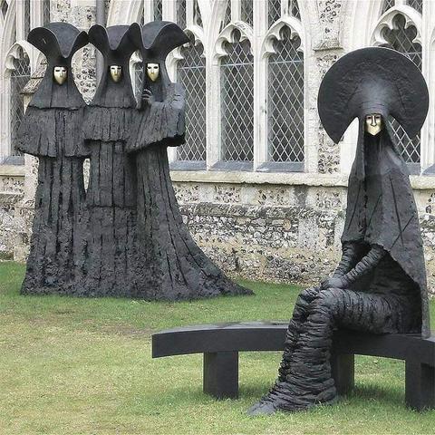 The figures are made of black iron with pale, white faces. They are sculped as if wearing black robes that invoke vaguely religious overtones, with elaborate head pieces and a severe aesthetic.