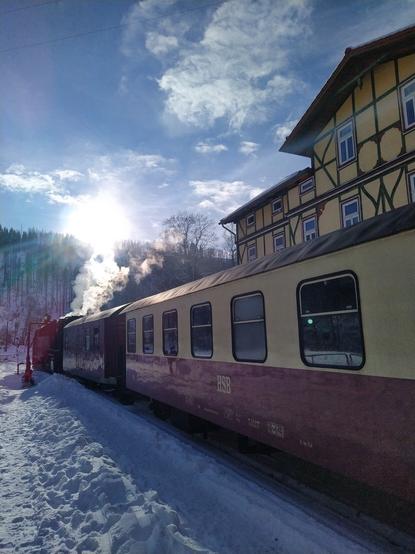 Ein Dampfzug mit beige-roten Wagen am verschneiten Bahnsteig. Im Hintergrund ein gelbes Fachwerk-Gebäude.